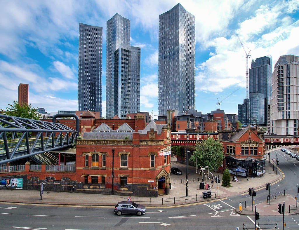 Historic brick building and elevated pedestrian bridge in front of a skyline of tall glass skyscrapers on a sunny day.
