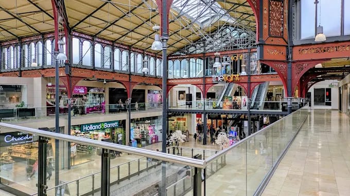 Interior of a Victorian-style shopping mall with glass ceiling and red decorative arches, storefronts on two levels with shoppers present.