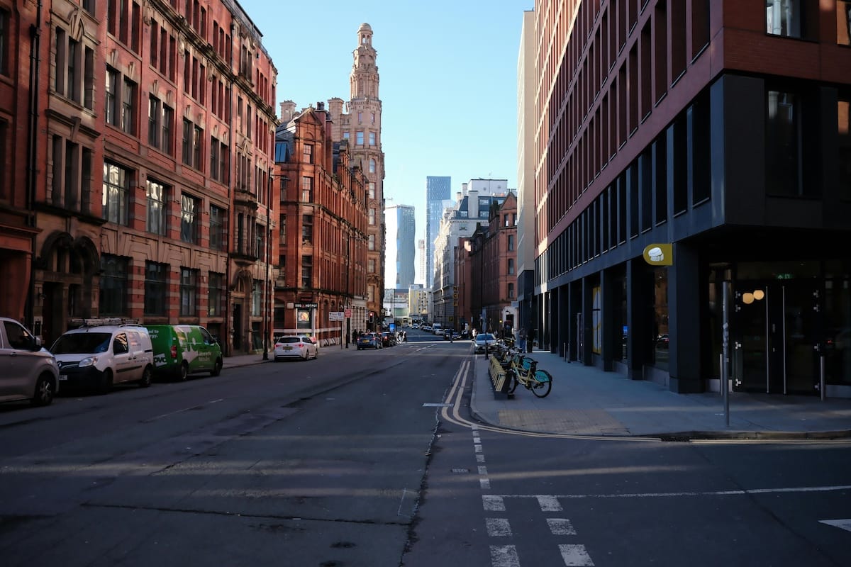 City street with red brick buildings on the left and a modern dark building on the right, bikes parked at the curb, and distant skyscrapers on the horizon.