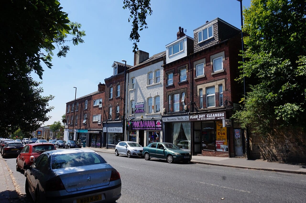Row of brick storefronts on a sunny street with parked cars and trees along the sidewalk.