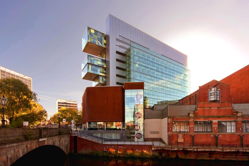 Modern glass and brick riverside building at sunset, with a tall glass atrium and red brick annex beside a canal.