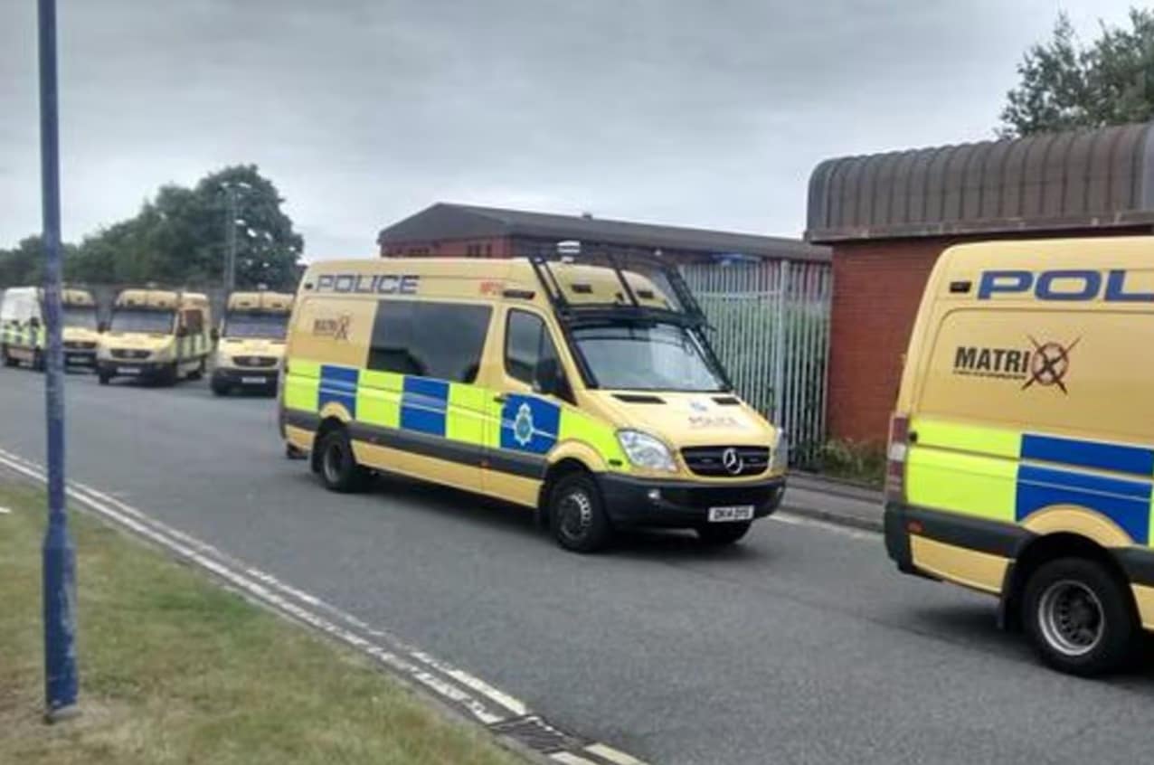 Line of yellow police vans with blue reflective chevrons parked along a street beside a fence and brick building on a cloudy day.