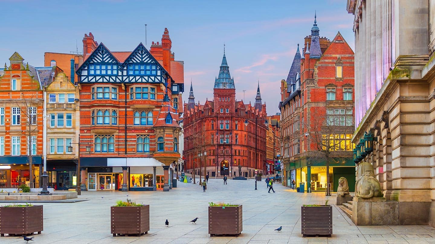 Historic red-brick city square with ornate gabled buildings, shopfronts, and pigeons on a paved plaza.
