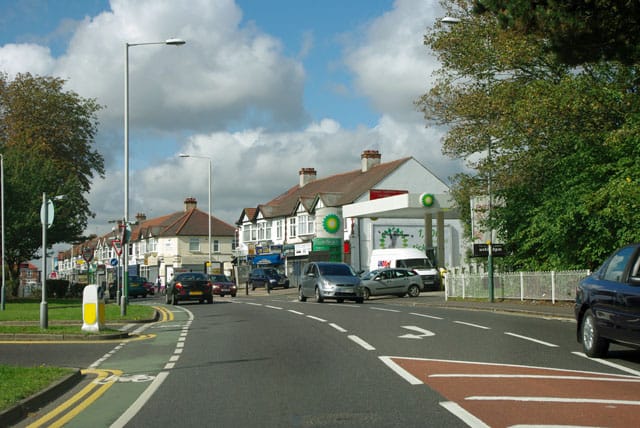 Town street with a BP gas station and row of shops along a two-lane road, cars passing by under a blue, partly cloudy sky.