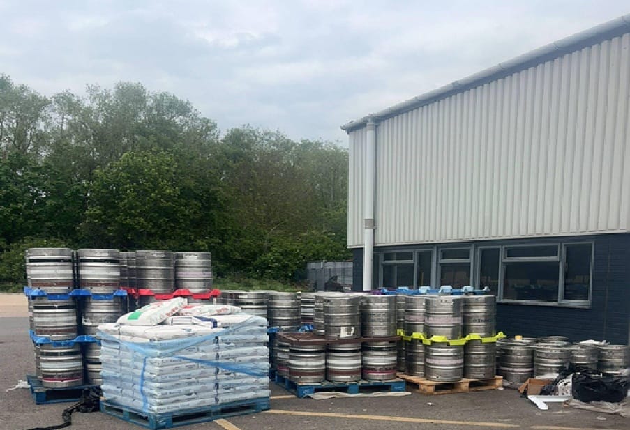 Outdoor storage area with stacked metal beer kegs on pallets beside a light industrial building and trees in the background.